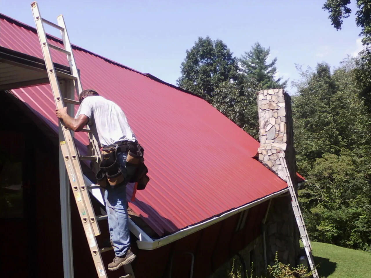 Roofer on a ladder beside a red metal roof and stone chimney — Soffit & Fascia Repair by Roofing Brothers in Cade